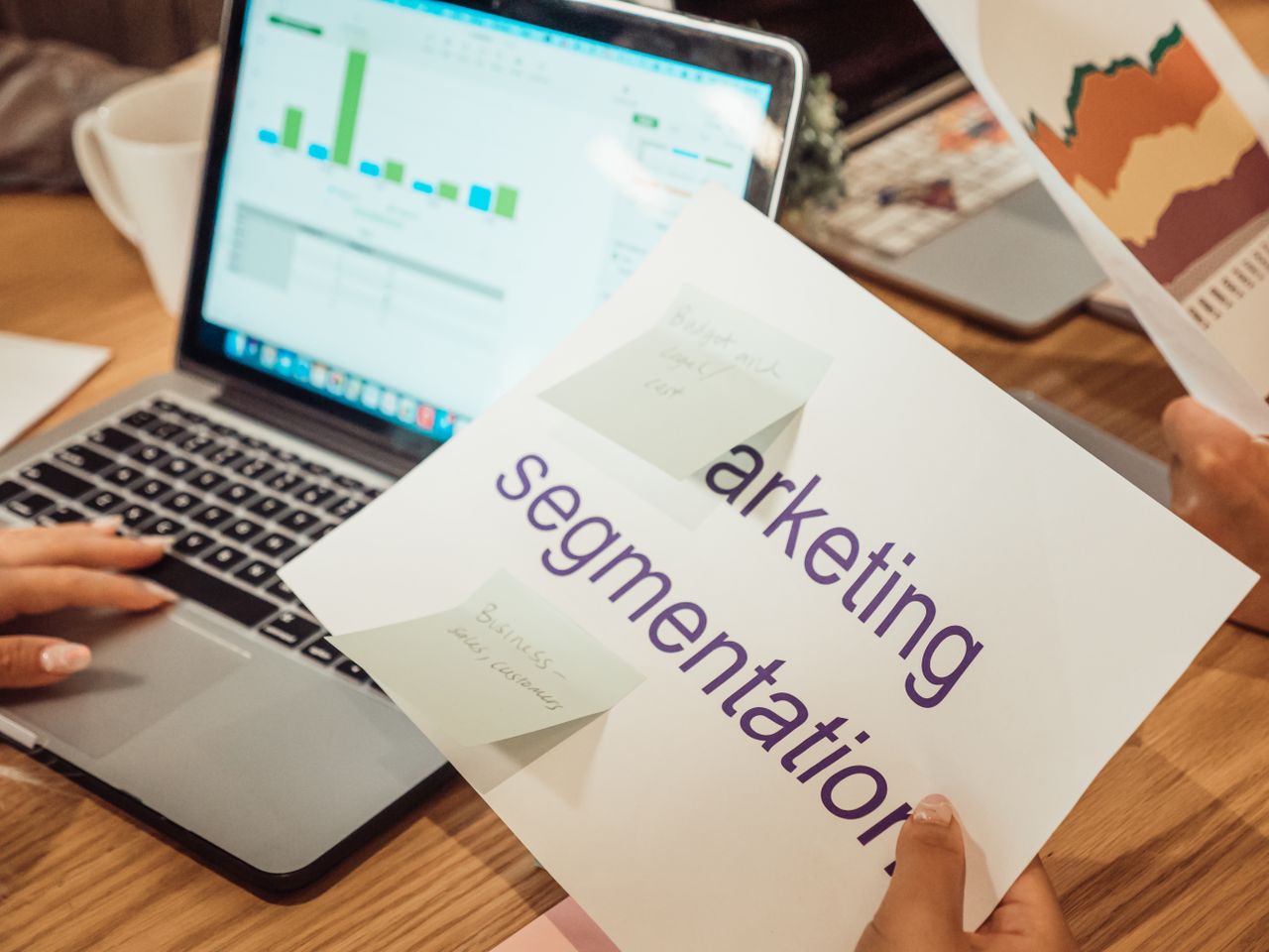 A contemporary workspace where hands review marketing data on a laptop alongside printed documents.